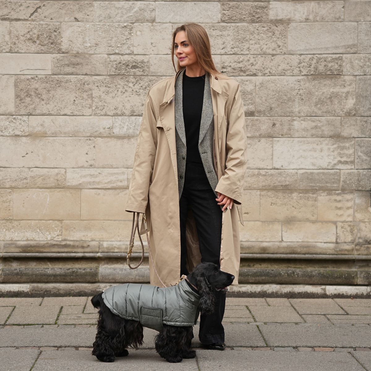 Woman walking a black terrier in a grey reflective coat past a stone wall