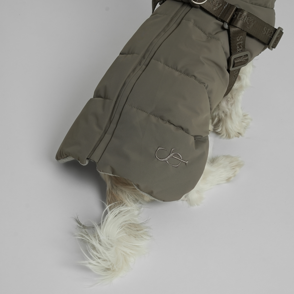 The moss green quilted coat with harness on a white dog, seen from behind